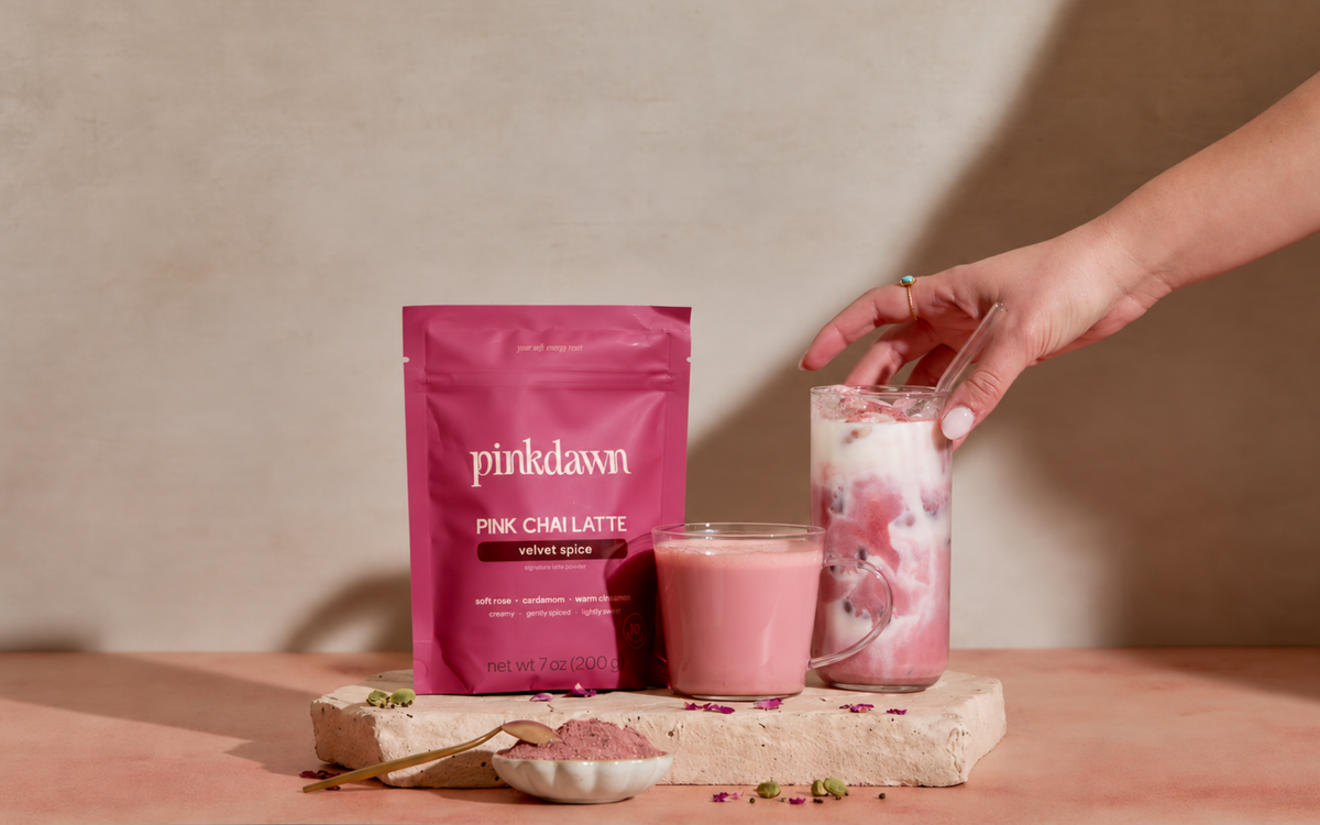 A hand holds a glass of pink chai latte next to a pink pouch of Pinkdawn Pink Chai Latte velvet spice on a stone slab, with a small bowl of powder and a spoon on a pink surface against a beige wall.