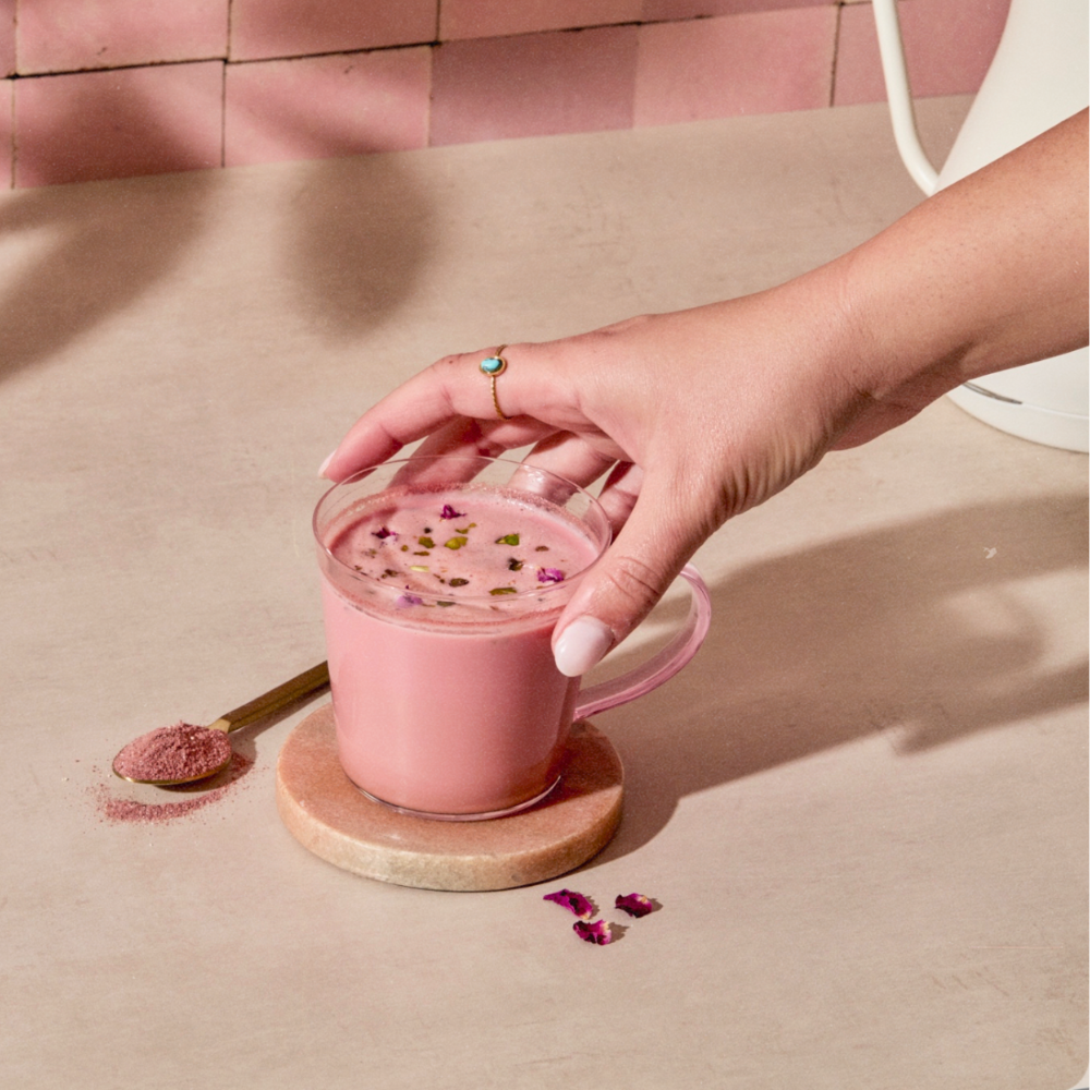 A hand with pink nail polish holds a clear glass mug filled with a pink chai beverage topped with dried flower petals, resting on a round marble coaster. A small spoon of matching pink powder lies nearby on a beige surface, with a pink tiled background.