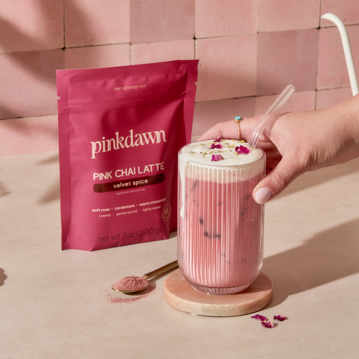 Iced PinkDawn pink chai latte with cold foam and rose petals, served in a glass (not included), beside the Velvet Spice pouch and a scoop of powder.