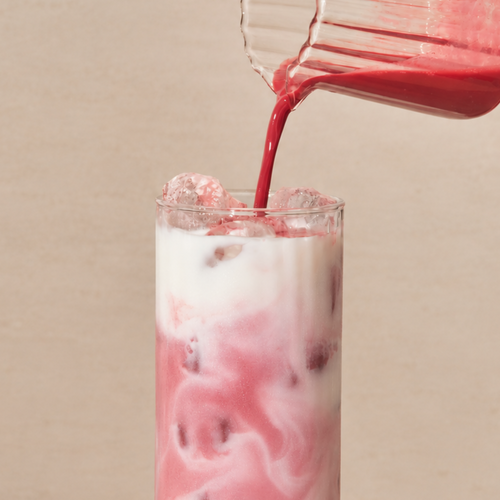A close-up of an iced pink chai beverage being poured from a glass pitcher into a tall clear glass filled with ice and a white liquid, against a warm beige background.